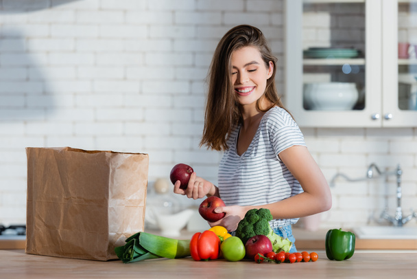mujer positiva sosteniendo manzanas jugosas cerca de bolsa de papel y verduras en la mesa de la cocina - Foto, Imagen