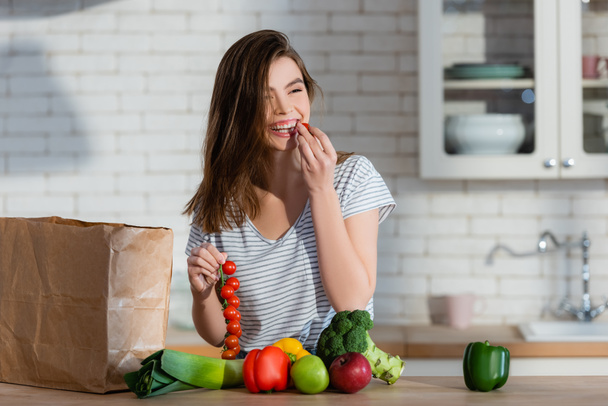 mujer riendo comiendo tomate cereza cerca de manzanas y verduras frescas en la cocina - Foto, Imagen