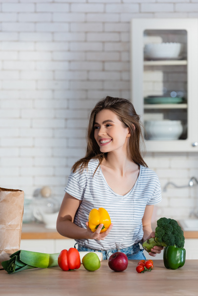 donna felice che distoglie lo sguardo tenendo in mano broccoli e peperoni in cucina - Foto, immagini
