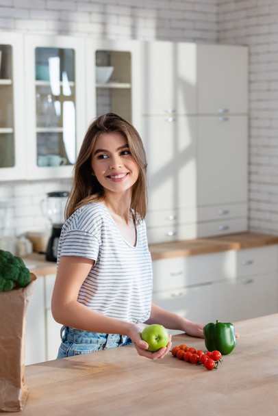 улыбающаяся женщина смотрит в сторону, держа яблоко рядом со свежими овощами на кухне - Фото, изображение