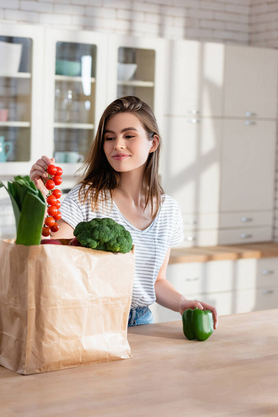 счастливая женщина держит ветку помидоров черри возле бумажного пакета со свежими овощами - Фото, изображение
