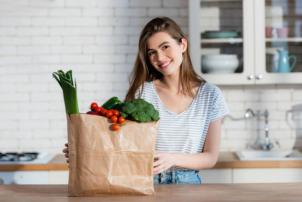 mulher alegre sorrindo para câmera perto de saco de papel com legumes na cozinha - Foto, Imagem