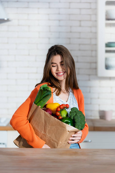 joyful woman holding paper bag with fresh vegetables in kitchen - Photo, Image