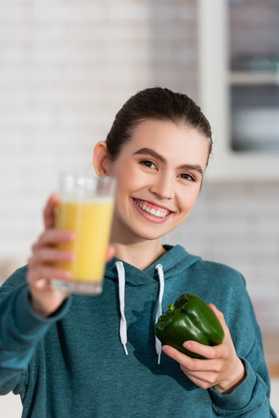 happy woman holding glass of smoothie and bell pepper while looking at camera on blurred foreground - Photo, Image