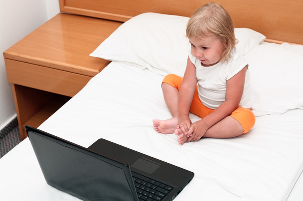 A small child is sitting with a laptop on the bed and looking at screen - 写真・画像