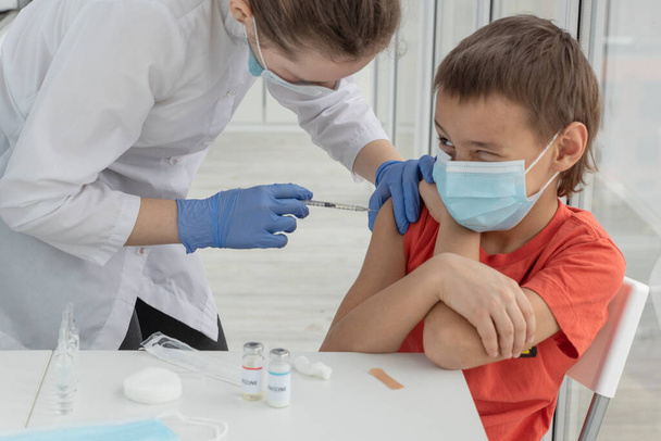 el muchacho divertido en la máscara médica está lokking con un ojo en doctor femenino que está haciendo la inyección de la vacuna en el brazo del muchacho. Emociones infantiles durante la vacunación - Foto, imagen