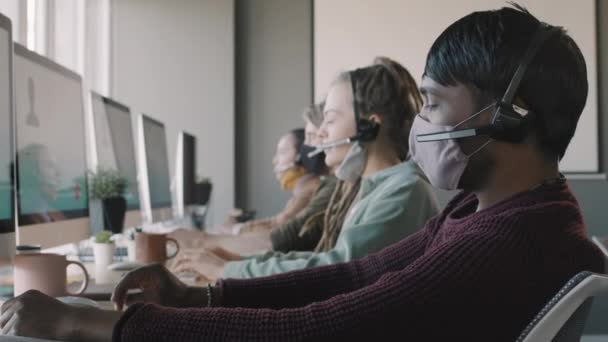 PAN oldalnézet lövés többnemzetiségű fiatalok headsetek és maszkok ül számítógép monitorok call center és értékesítési vagy beszél az ügyfelekkel - Felvétel, videó