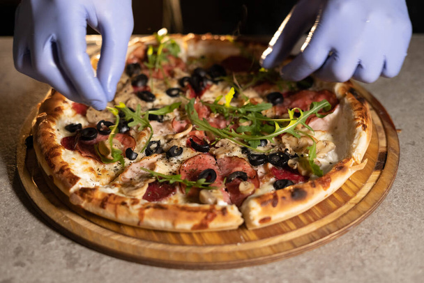 Руки шеф-повара в синих перчатках украшают готовую горячую пиццу листьями рукколы. Пеку крупным планом. Копирование пространства. - Фото, изображение