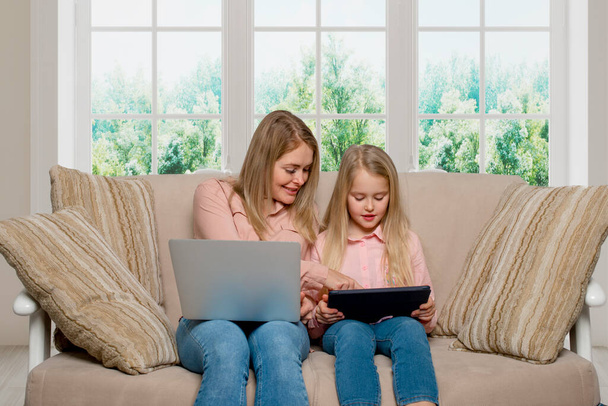 Egy kép egy anyáról és lányáról, hosszú hajú szőkékkel, rózsaszín ingben és farmerban, ahogy otthon ülnek a kanapén egy laptoppal és egy tablettával. A lánya utánozza anya viselkedését. - Fotó, kép