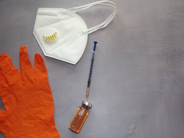 на сером фоне, вакцина, шприц, оранжевые медицинские маски и перчатки - Фото, изображение