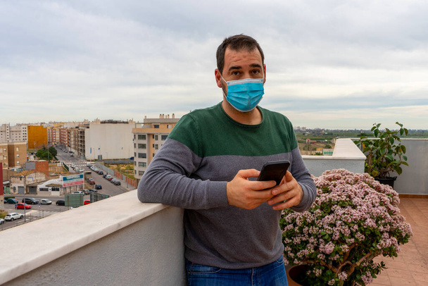 Ein bärtiger Mann in grauem und grünem Pullover mit Operationsmaske, um sich vor dem Coronavirus zu schützen, blickt besorgt auf sein Handy auf der Terrasse seines Hauses. Technologie und Gesundheitskonzept - Foto, Bild