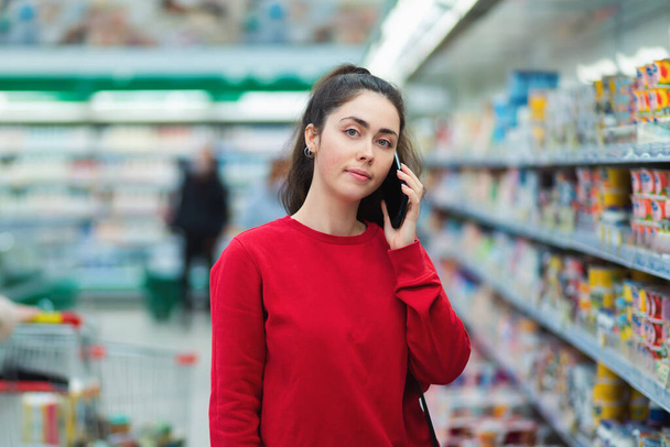 Portrét mladé ženy mluví po telefonu v supermarketu. Koncept komunikace a moderních technologií. - Fotografie, Obrázek