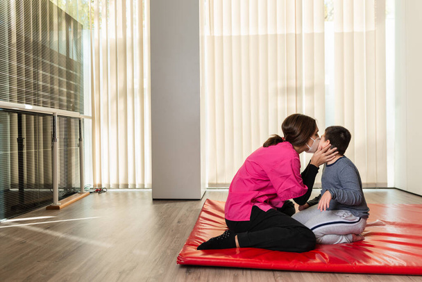 Ein behindertes Kind und ein Physiotherapeut küssen sich auf einer roten Matte und machen Übungen. Pandemieschutz - Foto, Bild