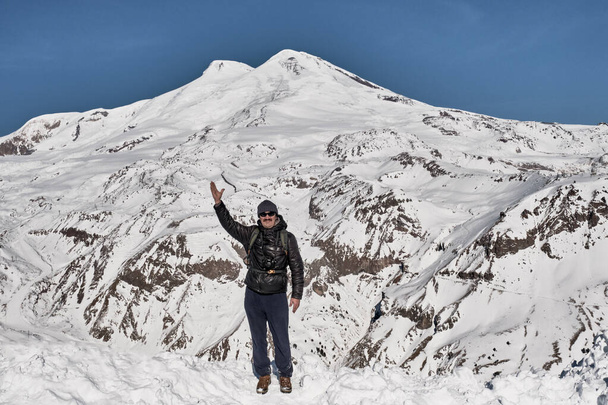 Zimowy krajobraz górski. Dojrzały mężczyzna z plecakiem w ciepłych ubraniach sportowych i okularach przeciwsłonecznych podniósł rękę, witając ją na tle dwóch szczytów góry Elbrus na Północnym Kaukazie, Rosja. - Zdjęcie, obraz