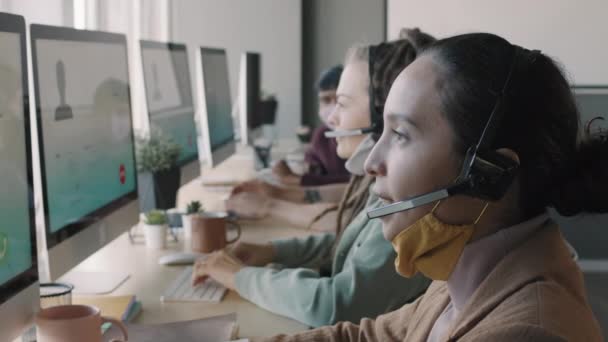 Slowmo puolella näkymä PAN nuorten Call Center agenttien kuulokkeet ja kasvonaamiot istuu ennen tietokoneen näyttöjä ja auttaa asiakkaita tai tehdä myyntiä - Materiaali, video