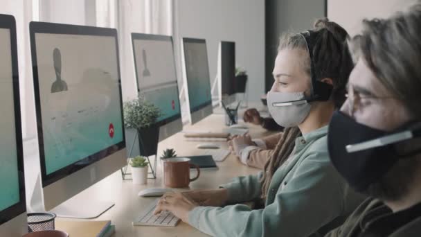 Zijaanzicht PAN van jongeren in gezichtsmaskers en headsets die voor computermonitoren zitten en klanten raadplegen bij audiogesprekken - Video
