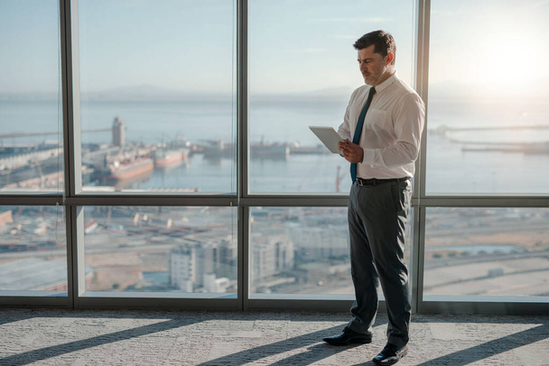 Зрілий бізнесмен в сорочці і краватці, використовуючи цифровий планшет, стоячи біля вікна в офісі з видом на місто
 - Фото, зображення