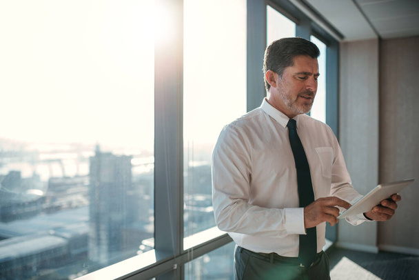 Idős üzletember, aki inget és nyakkendőt visel digitális táblagéppel, miközben egy ablakban áll, a városra néző irodában. - Fotó, kép