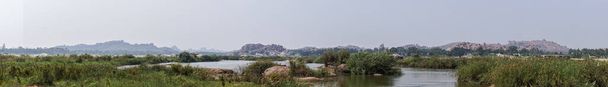 Anegundi, Karnataka, India - November 9, 2013: Wide Panorama shot over Gray Tungabhadra River with green covered islets and brown rock hills on horizon under light blue sky. - Φωτογραφία, εικόνα