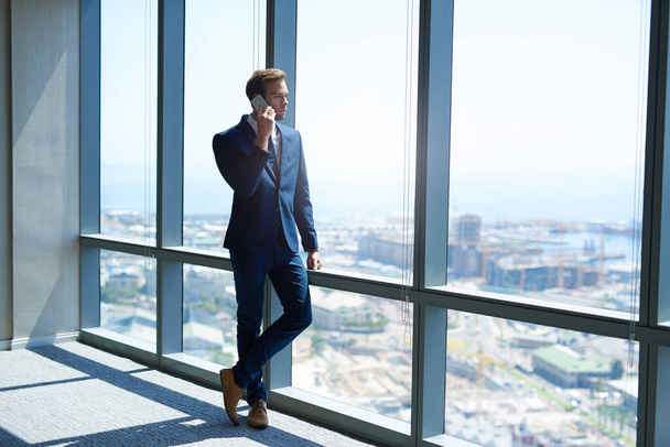 Foto completa de un joven hombre de negocios con estilo de pie junto a las grandes ventanas de una oficina moderna en lo alto de la planta superior, utilizando su teléfono mientras mira hacia la vista - Foto, Imagen
