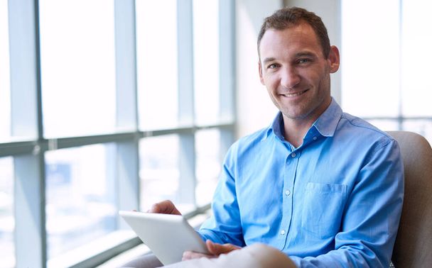Retrato de un hombre de negocios confiado sonriendo calurosamente a la cámara mientras está sentado en una oficina moderna y brillante y sosteniendo una tableta digital - Foto, imagen