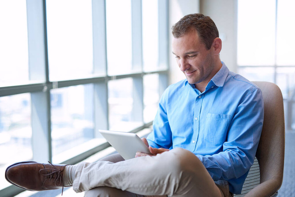 Uomo d'affari di mezza età seduto comodamente, utilizzando un tablet digitale in un luminoso ufficio moderno con ampie finestre - Foto, immagini