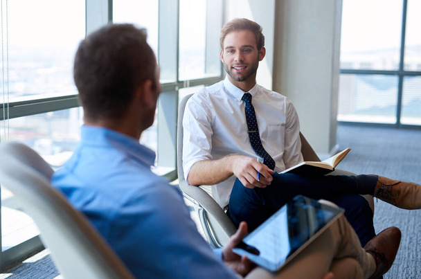 Νεαρός επιχειρηματίας κάθεται άνετα σε ένα ανοιχτό σύγχρονο γραφείο, χαμογελώντας ενώ έχει μια θετική συνομιλία με έναν συνάδελφο - Φωτογραφία, εικόνα