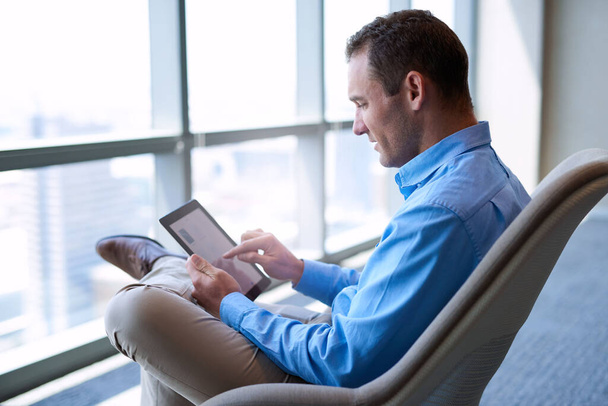 Üzletember ül a nagy ablakok előtt egy modern irodában, egy digitális tablet érintőképernyőjével. - Fotó, kép