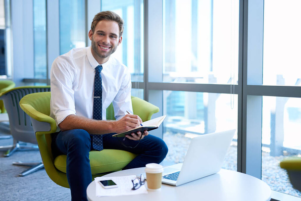 Portret van een jonge professional in een moderne bedrijfsruimte die aantekeningen maakt in zijn dagboek met zijn laptop voor hem open, en vol vertrouwen glimlacht naar de camera - Foto, afbeelding