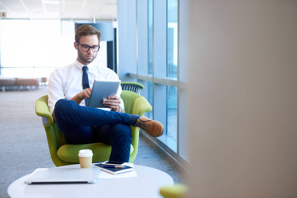 Elegante jovem empresário sentado confortavelmente em uma cadeira moderna em um espaço de escritório aberto, trabalhando em seu tablet digital  - Foto, Imagem