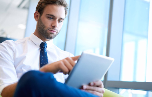 Vista de bajo ángulo de un joven hombre de negocios guapo con una corbata elegante, utilizando una tableta digital mientras está sentado junto a grandes ventanas  - Foto, Imagen