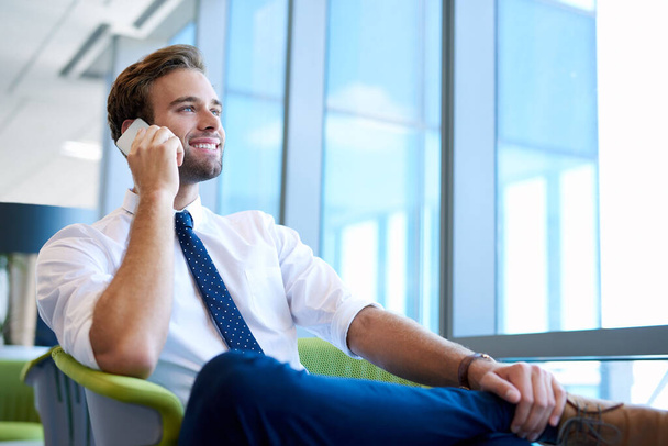 Low angle shot van een knappe ondernemer, zittend in een helder modern kantoor en breed glimlachend terwijl hij uit grote ramen kijkt en praat op zijn mobiele telefoon - Foto, afbeelding