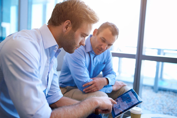 Zwei Führungskräfte sitzen in einem hellen Büroraum und suchen gemeinsam nach Informationen, indem sie den Bildschirm eines digitalen Tablets teilen.   - Foto, Bild