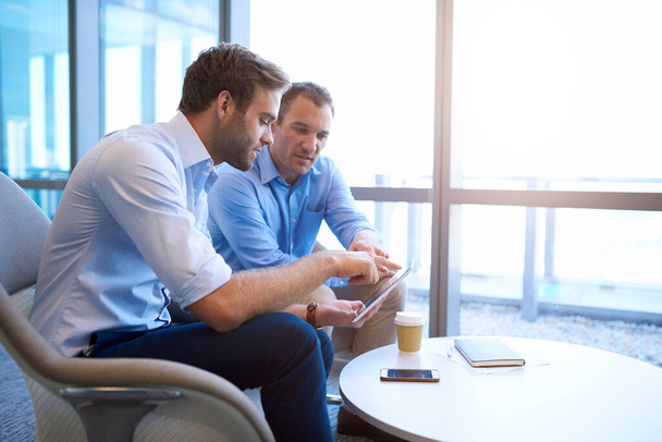 Два бізнесмени, що сидять у яскравому сучасному офісному просторі, використовують цифровий планшет у своїх обговореннях щодо нової співпраці
 - Фото, зображення