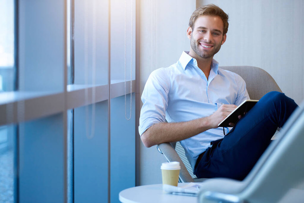 Portrét mladého podnikatele, který se sebevědomě usmívá, zatímco si zapisuje do deníku v moderní kanceláři se slunečními paprsky v okně vedle sebe - Fotografie, Obrázek