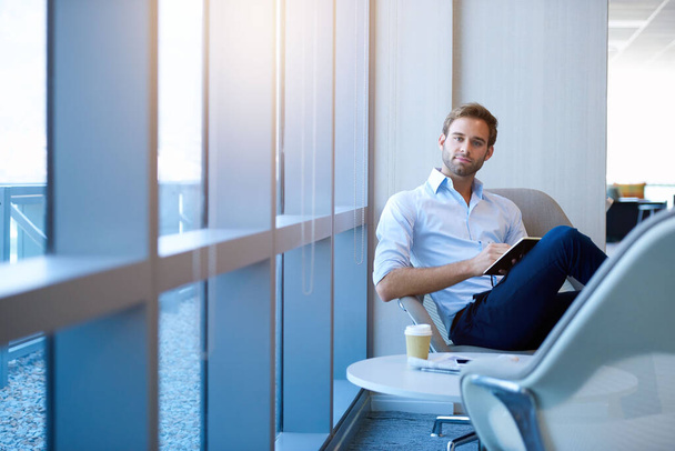 Portrét pozitivního mladého podnikatele sedícího v moderním firemním prostředí, dívajícího se do kamery a držícího svůj deník, s jemným slunečním světlem v oknech - Fotografie, Obrázek