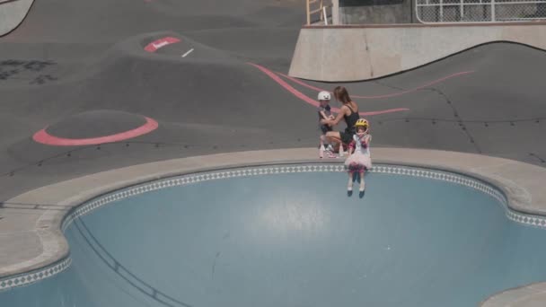 Amplia foto de una joven mujer caucásica usando ropa casual ayudando a su pequeña hija a ponerse equipo de seguridad mientras adolescente sentada en la parte superior de la rampa en el skatepark al aire libre y esperando - Imágenes, Vídeo