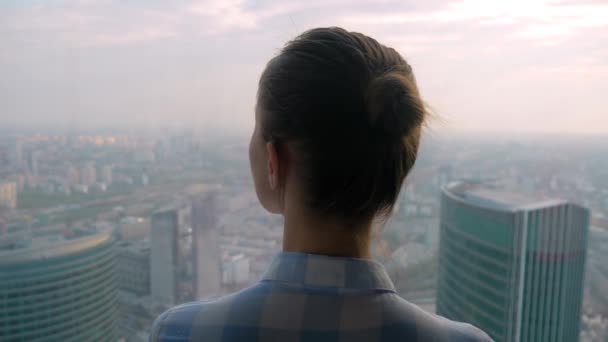 Achteraanzicht van pensive vrouw kijkend naar stadsgezicht door venster van wolkenkrabber - Video