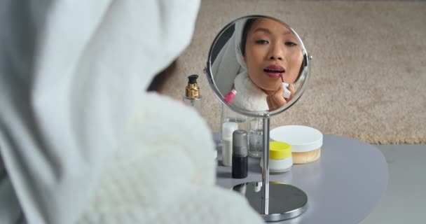 Heijastus nuori kaunis aasialainen korealainen nainen etninen tyttö peilissä pukeutuu valkoiseen kylpytakkiin ja pyyhe päähänsä maalaa huulensa huulipunalla tekee meikistä valmistautumisen tapahtumaan, kosmetiikkaan  - Materiaali, video