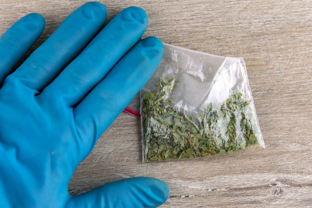 рука в перчатке прижимает к столу пакет синтетического дымящегося вещества, конопли, травы, марихуаны - Фото, изображение