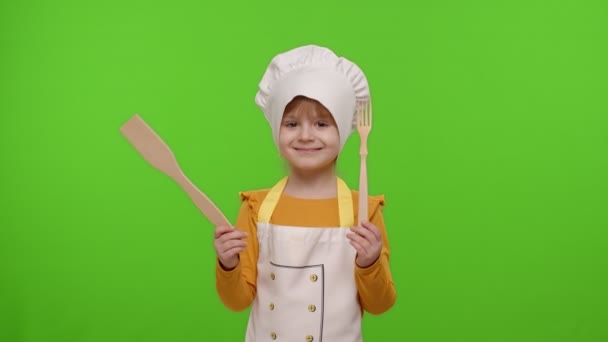 Enfant fille habillé comme chef cuisinier montrant fourchette et spatule en bois, souriant, hoche la tête en accord - Séquence, vidéo