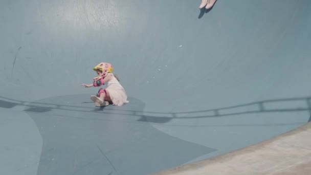 Ampio colpo di giovane donna caucasica indossa abiti casual seduto sulla cima della rampa in skatepark all'aperto e guardare le sue due figlie che indossano caschi di sicurezza scivolare giù tacco artificiale - Filmati, video