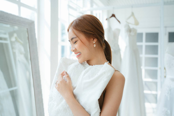 Eine Frau besucht Hochzeitsstudio für Hochzeitskleid Anprobe, sie heben das weiße Kleid, um über ihren Körper vor dem Spiegel zu setzen. - Foto, Bild