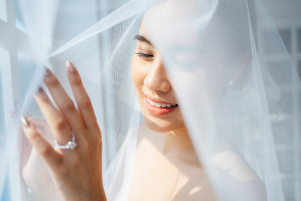 Pferdeschwanz blonde Haare Frau fertig verkleiden sich am Morgen ihres Hochzeitstages, steht neben dem Fenster, bedeckt sich mit weißem Schleier und berühren das Fenster, um ihren Ehering zu zeigen. - Foto, Bild