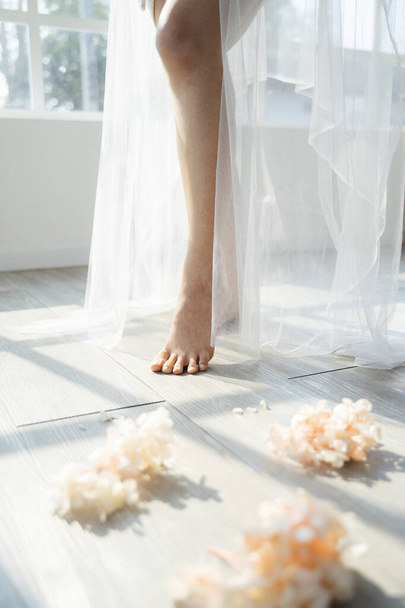 Περικοπή εικόνα των υστερήσεων της νύφης στέκεται με λευκό πέπλο ανάμεσα στο λουλούδι στο φως του ήλιου μέσα από το παράθυρο. - Φωτογραφία, εικόνα