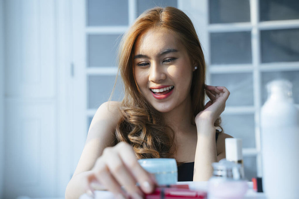 Το κυματιστό ξανθό κορίτσι διαλέγει το κόκκινο κραγιόν από το τραπέζι με χαμόγελο και κάνει το πρόσωπο πάπιας. - Φωτογραφία, εικόνα