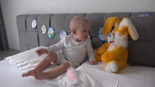 Aktywny chłopiec bawi się z dużym pluszowym królikiem w łóżku. Szczęśliwy dziecko pocałunek króliczek zabawka - Materiał filmowy, wideo
