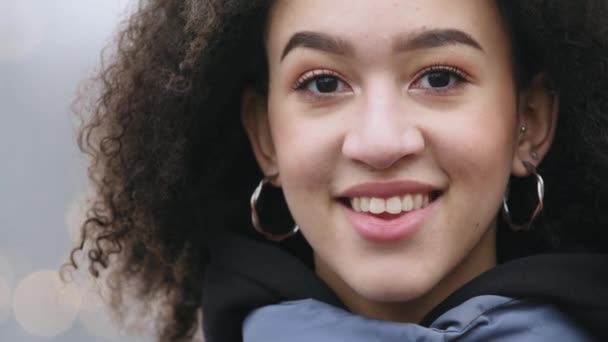 Vooraanzicht van Afro Amerikaans meisje met natuurlijke make-up draagt sieraden oorbellen staat op de achtergrond van de winter stad en wandelende mensen kijken naar camera glimlachend, close-up van vrouwelijke gezicht met donkere huid - Video