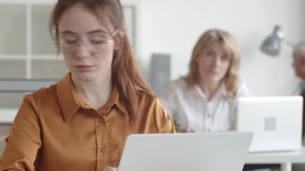 Kantelen close-up shot van jonge blanke vrouw zit in het kantoor met angstig gezicht, op zoek naar documenten en typen op laptop, en middelbare leeftijd supervisor kijken en friemelen met pen op de achtergrond - Video