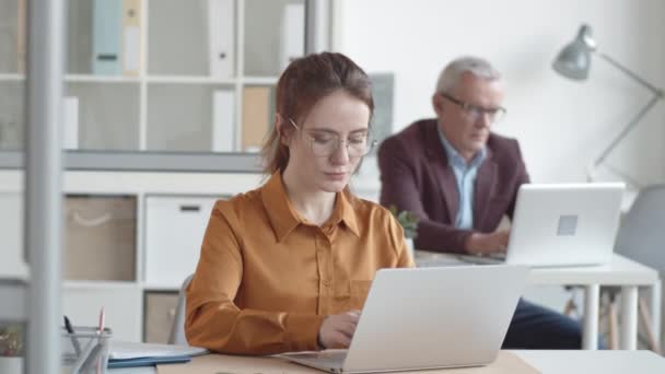 Waist-up shot van 20-iets blanke vrouw in bril en blouse zitten aan het bureau in het kantoor, typen op laptop met een lichte angstige glimlach, en middelbare leeftijd mannelijke baas werken op de achtergrond - Video